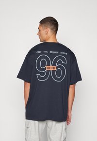 Námořnické bavlněné tričko s velkou bílou grafikou "96" na zádech, s oranžovými akcenty a tištěnými logy nahoře. Volný střih a kulatý výstřih.