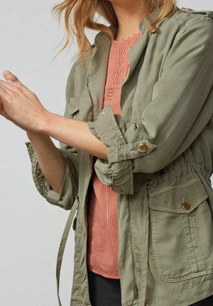 Žena si upravuje vyhrnuté rukávy olivově zelené bundy přes korálově texturovanou halenku a černé kalhoty, na jednobarevném pozadí.