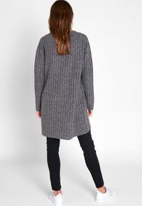 Žena s dlouhými vlasy, oblečená v dlouhém šedém pleteném svetru, černých kalhotách a bílých teniskách, stojící zády k bílé zdi.