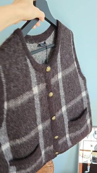 Brown and gray plaid vest made from soft wool, featuring gold buttons and two front pockets. Textured surface with a cozy appearance.