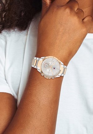 Sølv- og rosaguldur med en rund urskive, dekorative krystaller og et metalarmbånd, båret på et håndled mod en hvid skjorte baggrund.