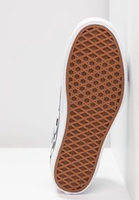 Semelle d'une basket blanche montrant une semelle extérieure en caoutchouc brun à motif gaufré avec un design géométrique antidérapant.