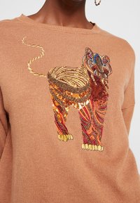 Pull en tricot marron présentant une broderie de lion colorée et pailletée aux motifs complexes, avec des accents dorés sur les pattes et la queue du lion.