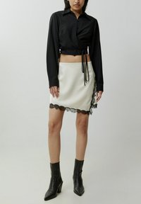 Schwarzes, langärmliges Cropped-Shirt aus strukturiertem Stoff, kombiniert mit einem weißen Minirock mit schwarzem Spitzenbesatz am unteren Rand und stylisch mit schwarzen Knöchelstiefeln.