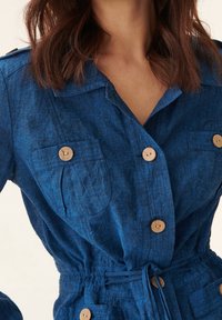 Blå linneklänning med knäppning fram, två bröstfickor och en markerad midja med dragsko. Texturerat tyg och träknappar.