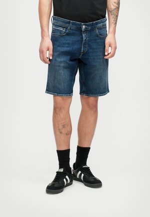 Bărbat îmbrăcat cu shorts din denim de culoare albastru închis, șosete negre și teniși negri cu dungi albe, stând în fața unui fundal deschis simplu.