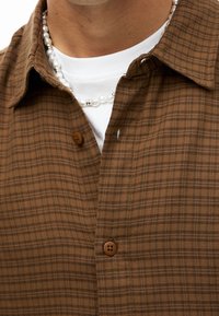 Camisa a cuadros marrón hecha de tela texturizada, con cierre frontal de botones marrones. Se lleva sobre una camisa blanca; se ve un collar de perlas.