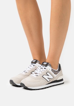 Pieds portant des baskets New Balance beiges avec des lacets blancs et des accents à imprimé léopard sur un fond blanc.