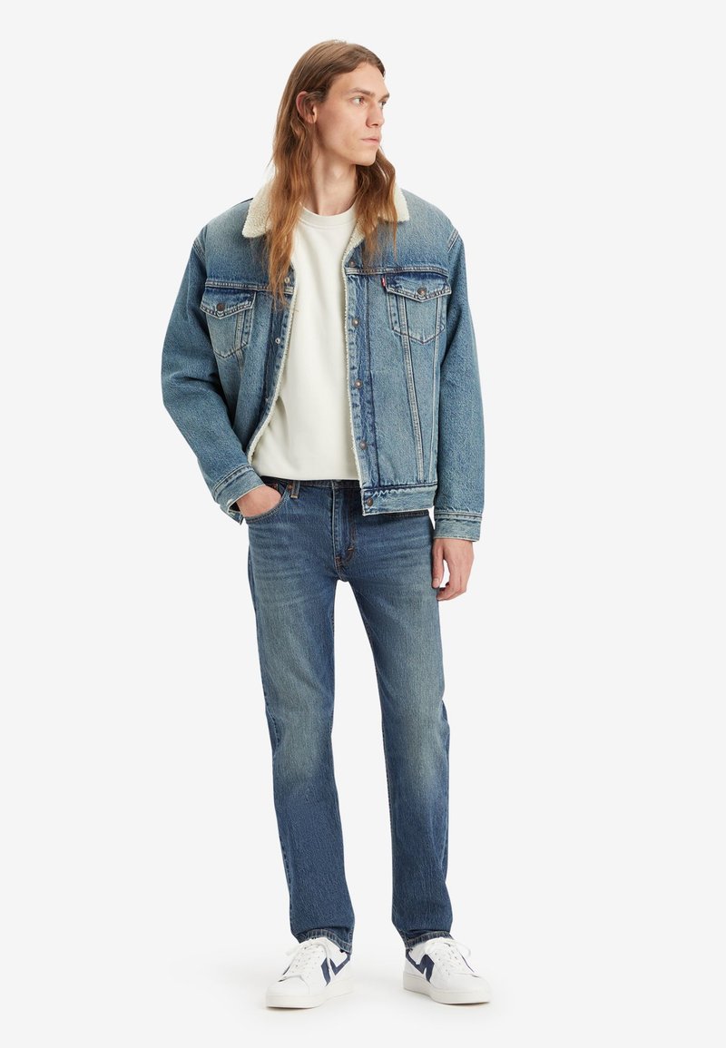 Jeansjacke mit Schaffellkragen, kombiniert mit einem weißen Sweatshirt und blauen Jeans. Das Model trägt weiße Sneakers mit marineblauen Akzenten.