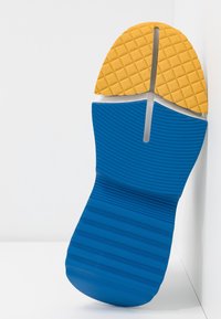 Sula av en blå och gul sportskor med texturerade greppmönster, lutande mot en vit vägg på en vit yta.