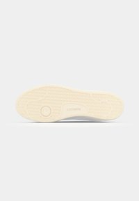 White sneaker with a patterned rubber sole. Features a textured upper and Lacoste branding embossed on the side of the sole.