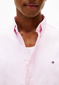 Camicia button-up rosa chiaro con colletto leggermente rialzato e logo ricamato sul petto. Il tessuto appare texturizzato e informale.