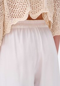 Beige, gebreide top met gekartelde randen, gedragen over witte, elastische broek met taille. Focus op de textuur en het lichte, vloeiende materiaal van de broek.