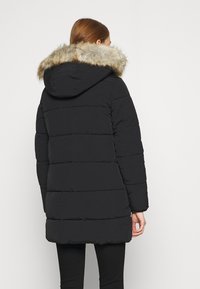 Žena s červenými vlasy, oblečená v černém prošívaném zimním kabátu s kapucí lemovanou kožešinou, zobrazená zezadu na jednoduchém pozadí.