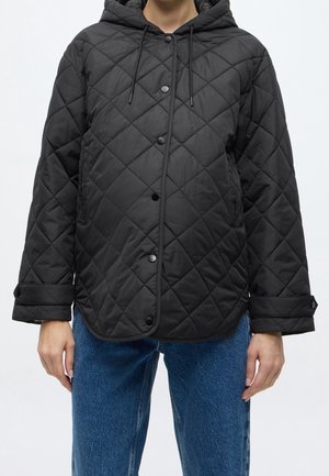 Person trägt eine schwarze gesteppte Jacke mit Kapuze, Druckknöpfen und verstellbaren Bündchen, kombiniert mit blauen Jeans vor einem einfarbigen Hintergrund.