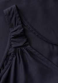 Ciemnoniebieska tkanina bluzki z marszczonym wykończeniem na lewym ramiączku, gładka struktura i staranne przeszycia wzdłuż krawędzi. Lekki materiał.