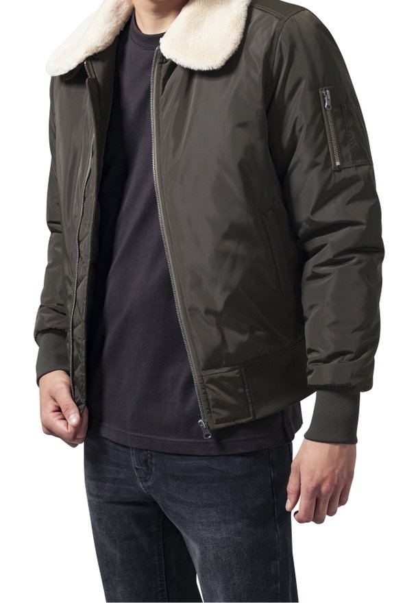 Homme portant une veste bomber vert foncé avec un col en polaire blanc, une chemise noire et un jean foncé, debout avec les mains détendues le long du corps.