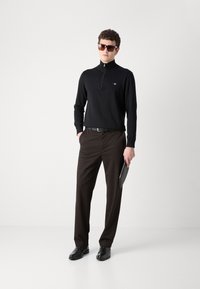 Crna majica dugih rukava sa zatvaračem i smeđe hlače. Ova odjeća upotpunjena je crnim cipelama i sunčanim naočalama, sa elegantnim, modernim dizajnom.