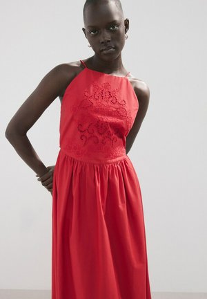 Junge Frau in einem roten ärmellosen Kleid mit Blumenstickerei auf der Brust steht vor einem schlichten hellen Hintergrund.