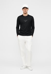 Mann mit weißer Baseballkappe, schwarzem Langarmshirt mit Logo, weißer Hose und weißen Turnschuhen, stehend mit den Händen in den Taschen.