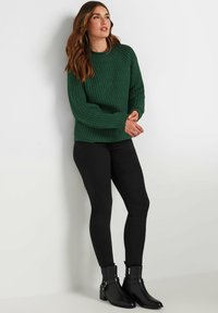 Chunky groene gebreide trui met textuurpatroon en lange mouwen, gecombineerd met zwarte nauwsluitende leggings en zwarte enkellaarzen met hardwareaccenten.