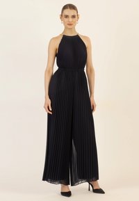 Combinaison plissée bleu marine avec un col halter, une taille élastique et des jambes larges. Présente un détail en chaîne au niveau du col et est portée avec des talons noirs.