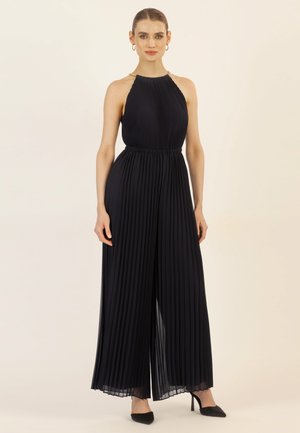 Marineblauwe geplooide jumpsuit met halternek, elastische tailleband en wijde benen. Heeft een kettingdetail op de halslijn en wordt gedragen met zwarte hakken.