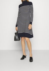 Nainen, jolla on päällään tummansininen ja valkoinen raidallinen pitkähihainen mekko, mustat korkokengät ja pieni musta käsilaukku.