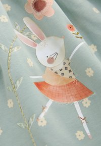 Lapin blanc portant une robe fleurie et une jupe orange, tenant une petite fleur rose, entouré de fleurs brodées sur un tissu bleu clair.