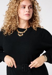 Zwarte gebreide trui met poffende mouwen, een ceintuurlijn en een gouden gelaagde ketting. Model draagt meerdere gouden ringen en armbanden.