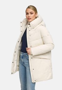 Femme portant une doudoune crème avec un col montant, un haut marine et un jean bleu clair sur un fond uni.