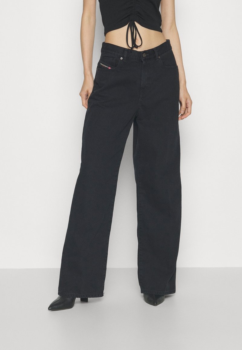 Personne portant un jean noir taille haute à jambes larges et un crop top noir sans manches, debout devant un fond blanc uni.