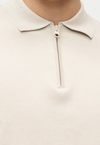 Polo beige en maille avec une courte fermeture éclair à l'avant et un col large et doux sur le torse supérieur d'une personne.