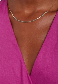 Gouden ketting met turquoise, witte en bruine kralen in een gebogen ontwerp, gedragen tegen een diepe V-halslijn van fuchsia stof.