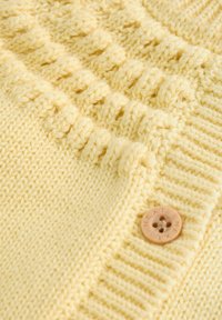 Knitana rumena jopica v rumeni barvi z teksturno šivano vzorčasto površino, rebrastimi detajli in lesenim gumbom z vgravirano besedo "NEXT".