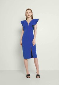 Vestido midi azul-rei com mangas flutuantes, decote em V e uma fenda frontal. Textura suave, design justo, combinado com sandálias de salto preto.