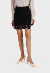 Minifalda de encaje negro con patrones florales y dobladillo de forma festoneada, combinada con unos tacones destalonados de punta fina en rosa pálido y tejido suave.