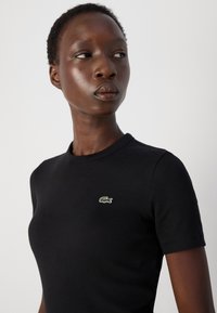 Juodas gumbas t-shirt su trumpomis rankovėmis ir apvalia iškirpte. Ant krūtinės yra mažas žalias ir raudonas išsiuvinėjimas krokodilo logotipo.