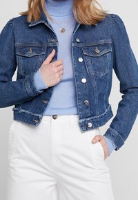 Kvinna i en croppad denimjacka över en ljusblå polotröja och vita högmidjade byxor, med ena handen i fickan och den andra hållande i jackans slag.