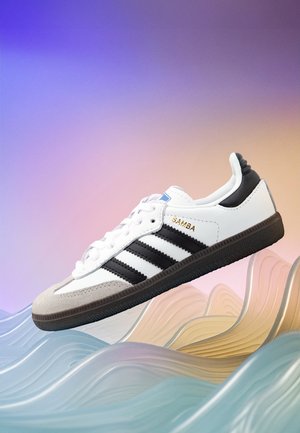 Fehér sneaker fekete csíkokkal, szürke velúr orral és texturált fekete talppal. Fókuszban a fűzők, a logó és a párnázott gallér.