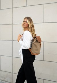 Blonde woman in white blouse and black pants standing with tan and brown backpack against tiled wall, looking over shoulder.