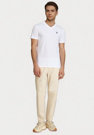 Wit T-shirt met V-hals, korte mouwen, zacht katoenen materiaal, gecombineerd met beige broek en beige sneakers, staand op een lichte achtergrond.