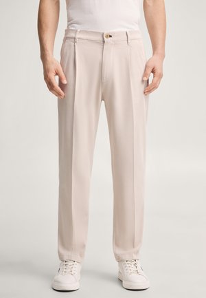 Homme portant un pantalon beige droit avec des plis, des baskets blanches et une chemise blanche, debout devant un fond uni.
