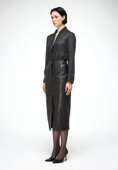 Žena nosí priesvitnú čiernu blúzku a čiernu koženú ceruzkovú sukňu s vysokým pásom a predným rozparkom, doplnenú čiernymi priesvitnými pančuchami a podpätkami.