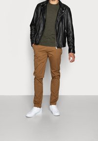 Veste de motard en cuir noir, t-shirt olive, pantalon marron fuselé et baskets blanches. Design simple avec des accents minimaux.