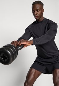 Fekete hosszú ujjú sportfelső és rövidnadrág, fekete kettlebellt fogva. Kiemelt hangsúly az izmos karokon és a definiált testalkaton, semleges háttér előtt.