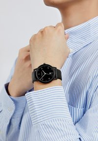 Reloj de hombre negro con una esfera redonda, marcas blancas y una correa de cuero negra, llevado en una mano con una camisa azul a rayas.