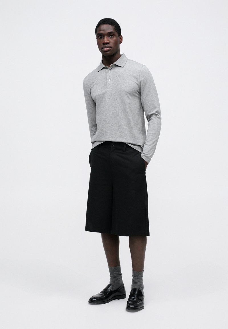 Polo grigio a maniche lunghe con colletto, abbinato a pantaloni corti neri fino al ginocchio, calze grigie e mocassini neri. Stile semplice e moderno.