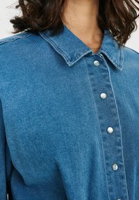 Denimskjorta med en klassisk krage, ljusblått tyg och silverfärgade tryckknappar på framsidan. Slät textur och strukturerad design.