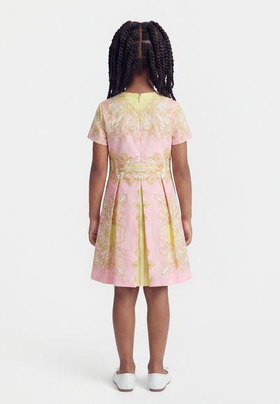 Νεαρή κοπέλα με πλεγμένα μαλλιά, φοράει φόρεμα με ροζ και κίτρινο μοτίβο και άσπρα παπούτσια, στέκεται γυρισμένη πλάτη σε λευκό φόντο.